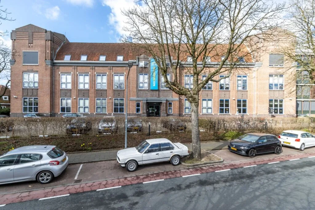 Vooraanzicht van een groot schoolgebouw aan de Lage Naarderweg met geparkeerde auto's en een boom op de voorgrond.